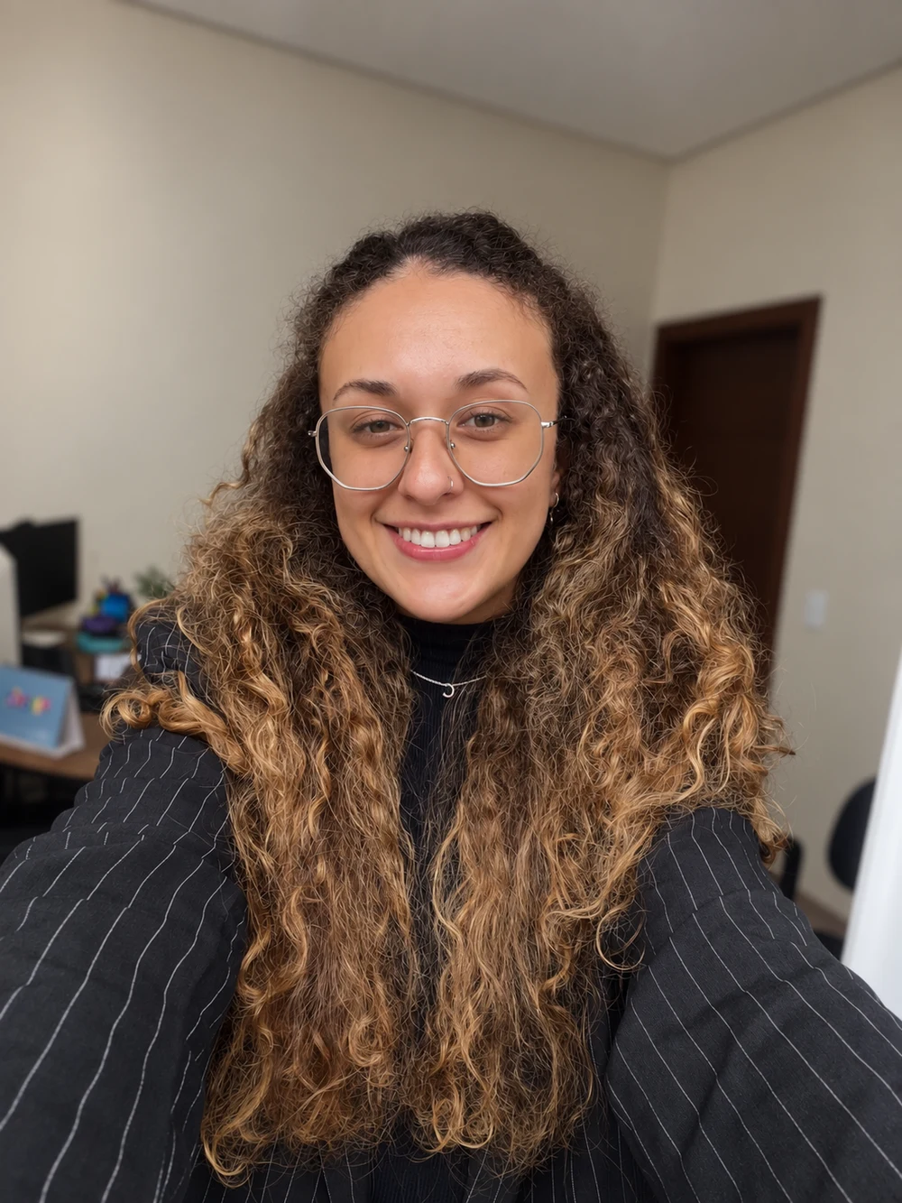 Camila Abreu sorrindo, com óculos e blazer de risca de giz, em seu ambiente de trabalho.